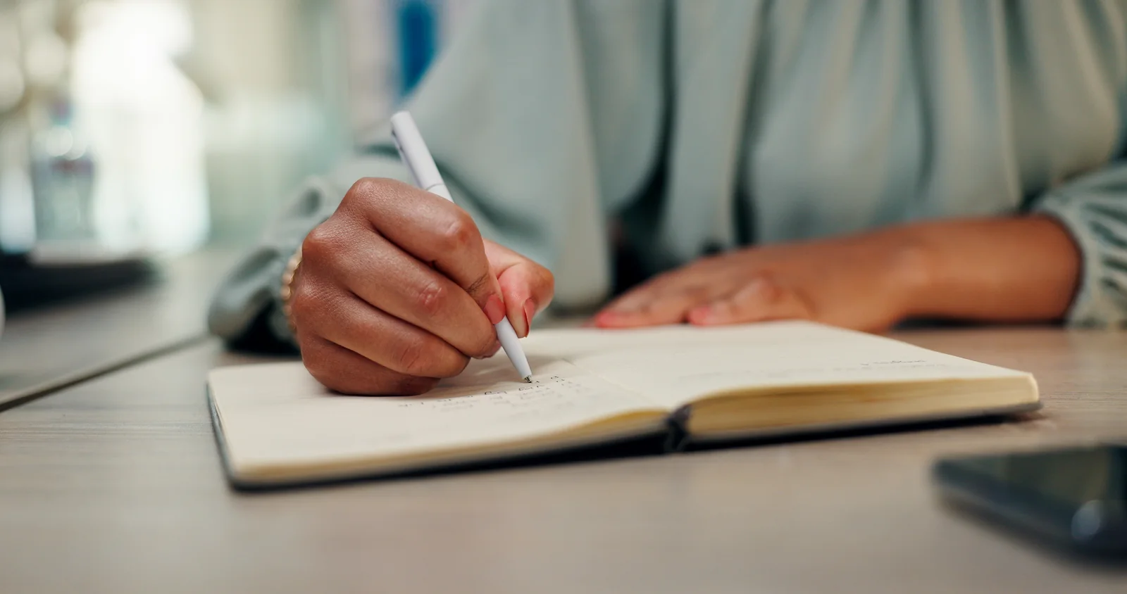 A woman’s hands writing in an open journal at a table.